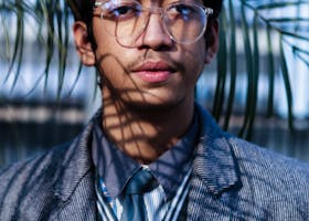 Portrait of a young man wearing eyeglasses and a suit in a natural setting with palm leaves.