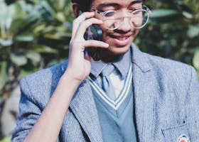 A young man wearing eyeglasses and a blazer smiling while talking on a smartphone outdoors.