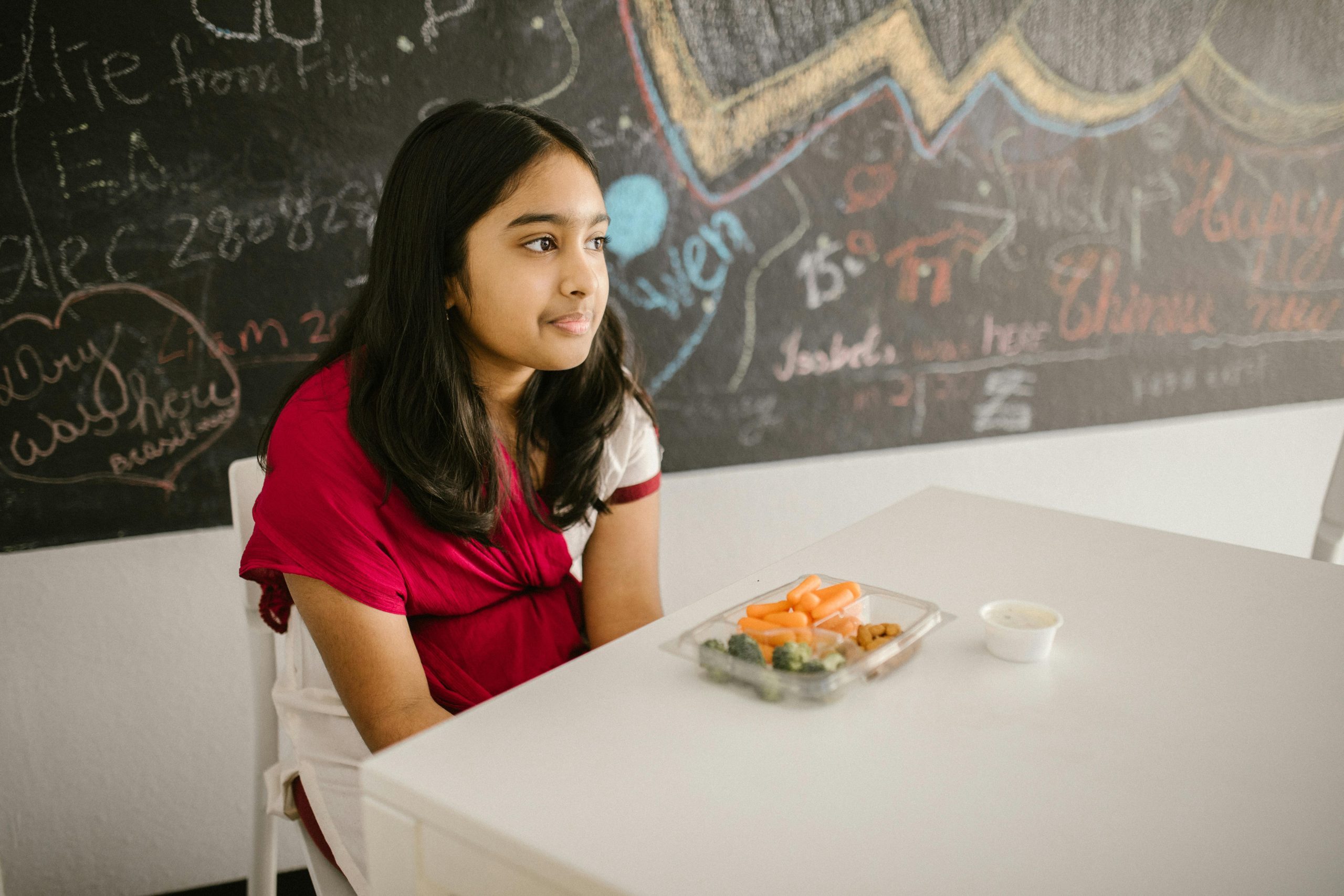 A young girl sits thoughtfully at a classroom table with a snack pack.